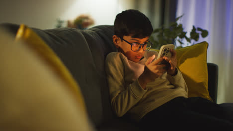 Young-Boy-Sitting-On-Sofa-In-Lounge-At-Home-Playing-Game-On-Mobile-Phone-At-Night-2